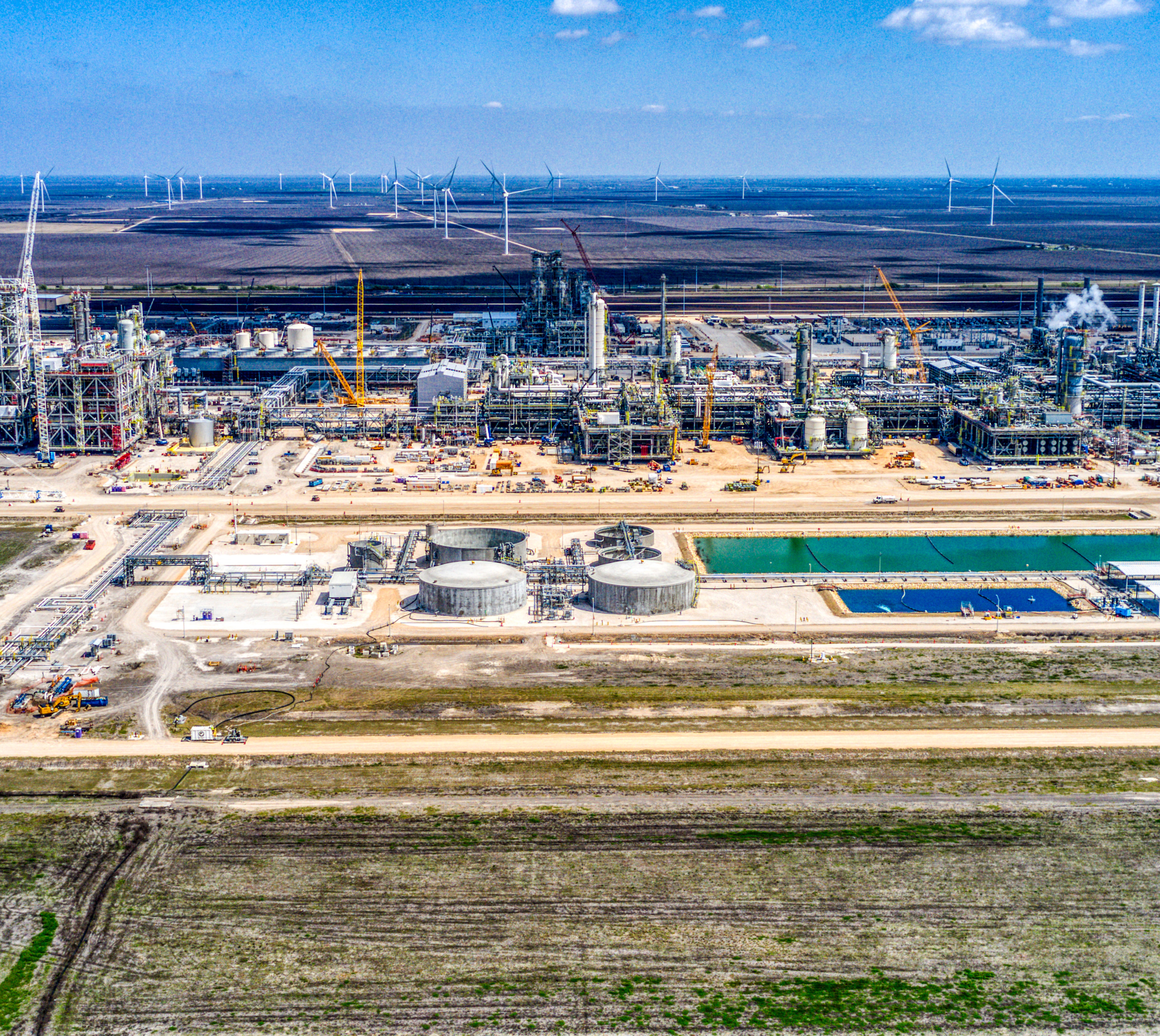 a refining site underconstruction. storage tanks for producs infront. to the right of the storage tanks is a water storage pool. behind the plant are turbines from a neighboring wind farm scattered across farm land.